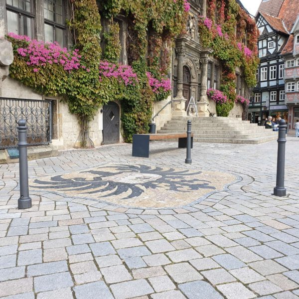 Wappen-Poller im steinernen Innenhof mit schwarzem Adler, umgeben von Pollern, Stufen, Bank und efeubewachsenem Gebäude mit Blumen.