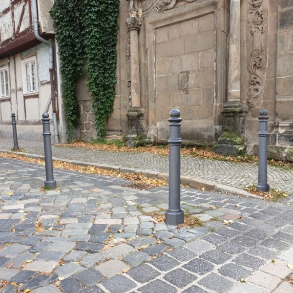 Vier graue Wappen-Poller aus Metall auf Kopfsteinpflaster neben historischem Gebäude mit Efeu und Herbstlaub auf dem Boden.
