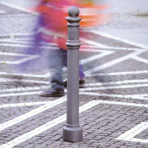 Grauer Wappen-Poller aus Metall auf Kopfsteinpflasterstraße mit weißen Markierungen, unscharfe Passanten im Hintergrund.