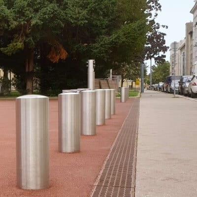 Mehrere CITY-Poller BERLIN XL Metallpoller stehen entlang eines roten Kieswegs am Gehweg, Bäume und parkende Autos im Hintergrund.