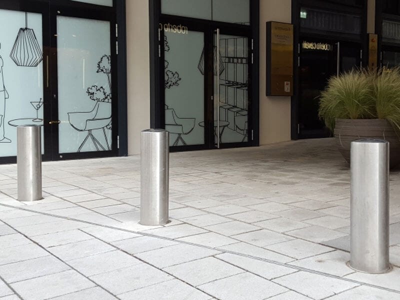 Drei CITY-Poller BERLIN XL aus Metall auf gepflastertem Gehweg vor Glastüren mit Fensterdekor, neben Pflanzkübeln mit Ziergras.