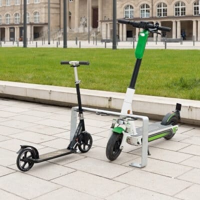 Person hält schwarzen Tretroller und grünen E-Scooter nebeneinander auf Gehweg, neben Rasenfläche und Gebäude in der Stadt.
