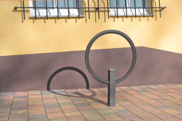 Runder CITY-Anlehnbügel GIRO Fahrradständer auf Fliesen vor gelb-brauner Wand mit vergitterten Fenstern und Schattenwurf.