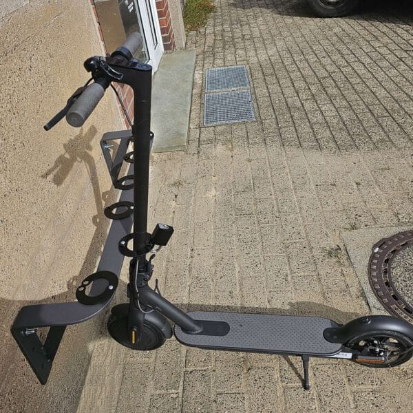Ein schwarzer E-Scooter steht vertikal im CITY-Scooter-Parker GILA an einer Wand, außen auf Pflaster neben einem Gullydeckel.