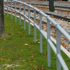 Metallzaun MORION Systemgeländer URBAN verläuft parallel zu Bahngleisen, umgeben von grünem Gras und fallenden Blättern.