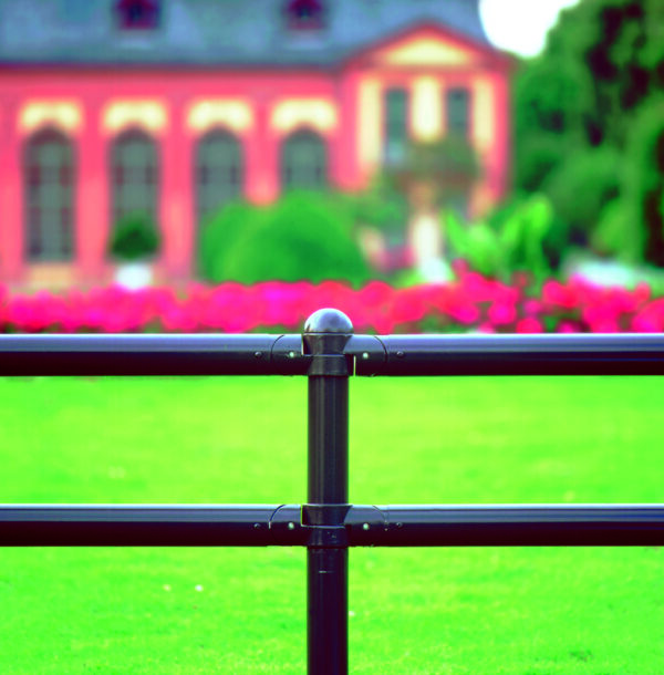 Nahaufnahme MORION Systemgeländer CLASSIQUE, schwarzer Metallzaun, grüner Rasen, rosa Blumen, unscharfes Gebäude im Hintergrund.