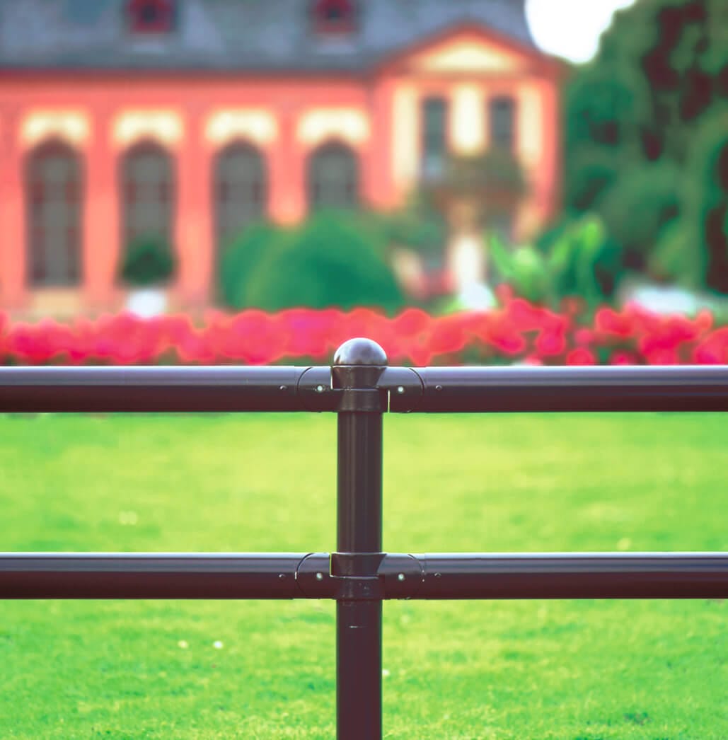 Nahaufnahme MORION Systemgeländer CLASSIQUE, schwarzer Metallzaun, grüner Rasen, rosa Blumen, unscharfes Gebäude im Hintergrund.