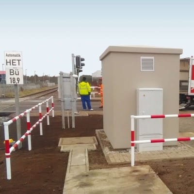 MORION Systemgeländer als Leitplanke neben Technikgehäuse an Bahnübergang, Arbeiter mit Warnweste bei Verkehrszeichen.