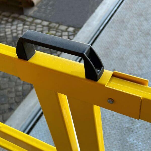 Nahaufnahme eines U-förmigen Stahlbügels mit schwarzem Griff an gelber Absperrung, Hintergrund: Gehweg und Beton.