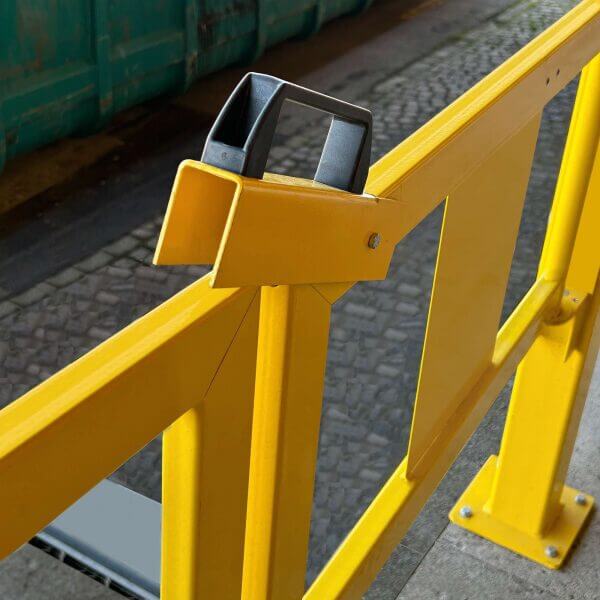 U-förmiger gelber Stahlriegel mit schwarzem und grauem Verschluss, montiert auf Kopfsteinpflaster neben grünem Container.