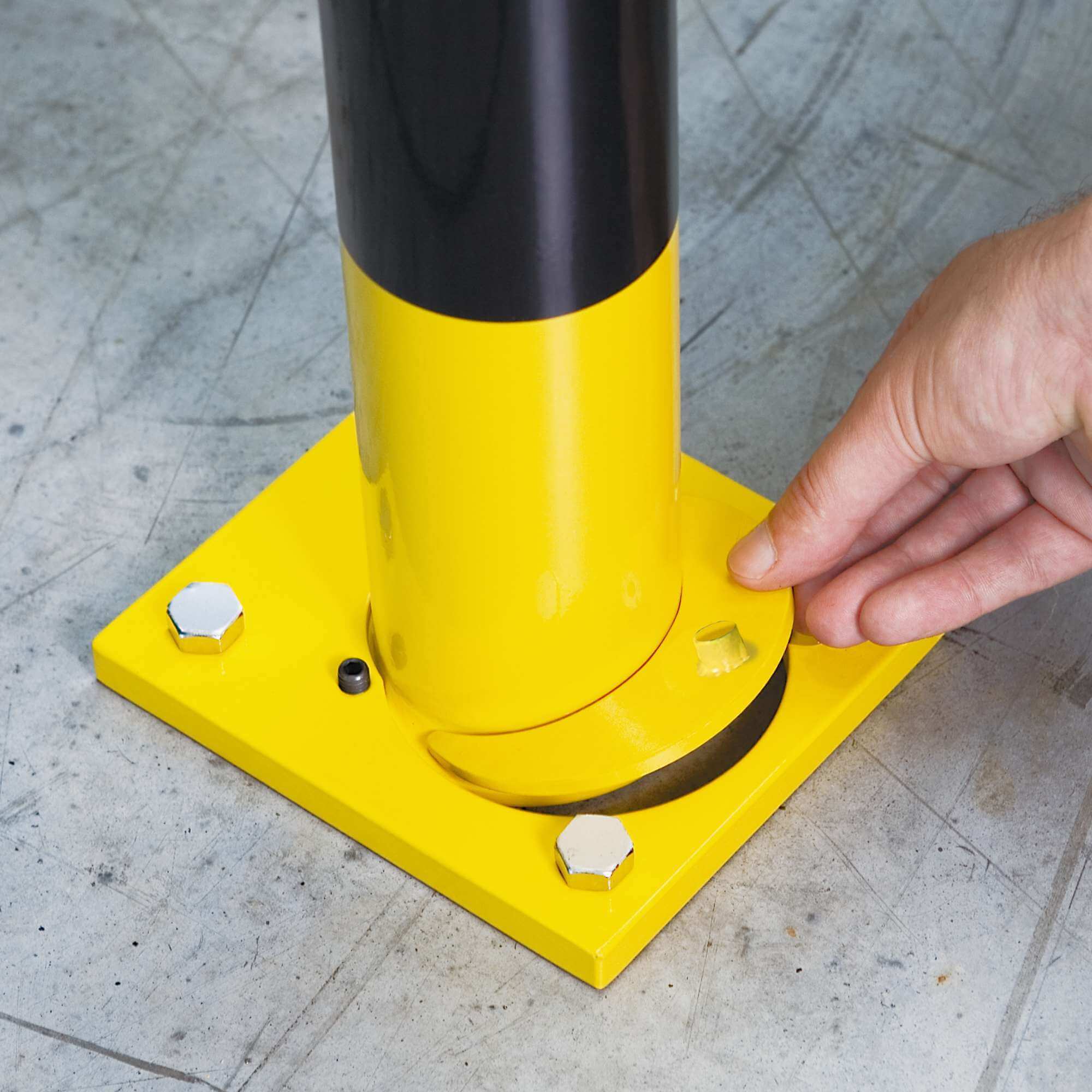 Hand hebt gelbe Grundplatte eines BLACK BULL Rammschutz-Bügels, schwarz-gelber Poller mit Bodenmontage, Betonboden, Schrauben.