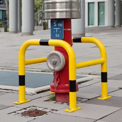 MORION Säulenschutz mit gelb-schwarzen Streifen schützt rote Hydranten mit Metallkappe und blauem Etikett auf grauen Fliesen.