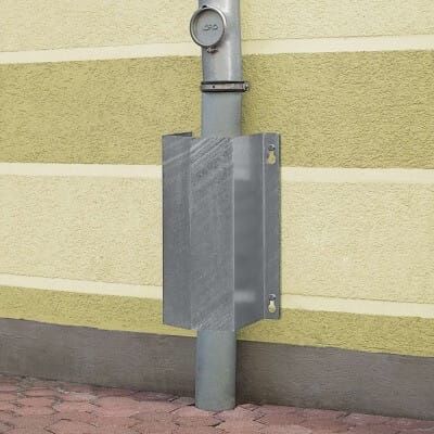 MORION Rohrschutz klein: Vertikaler Metallrohrschutz an Wand, teils von rechteckigem Metallschild bedeckt, über Pflaster.