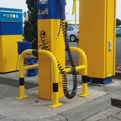 Gelb-blaue MORION Säulenschutz-Geländer umgeben eine Reifendruckluftpumpe an der Tankstelle mit Spiralschlauch und Schutzbügeln.