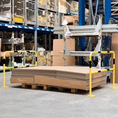 Flache Kartons gestapelt auf Holzpalette im Lager, umgeben von gelben MORION Schutzbügeln und Regalen mit Lagerware.