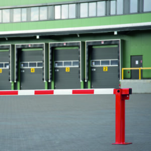Rote und weiße Systemschranke mit Gasdruckfeder blockiert Zufahrt vor vier geschlossenen Laderampen an Industriegebäude.