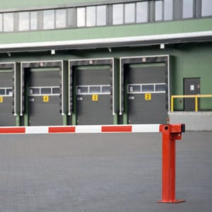 Rote und weiße Systemschranke mit Gasdruckfeder blockiert Zufahrt vor vier geschlossenen Laderampen an Industriegebäude.