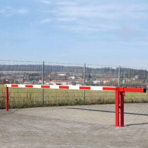 Rote und weiße COMPACT Systemschranke mit Gegengewicht sperrt gepflasterte Zufahrt mit Drahtzaun, Wiese und Wolkenhimmel.