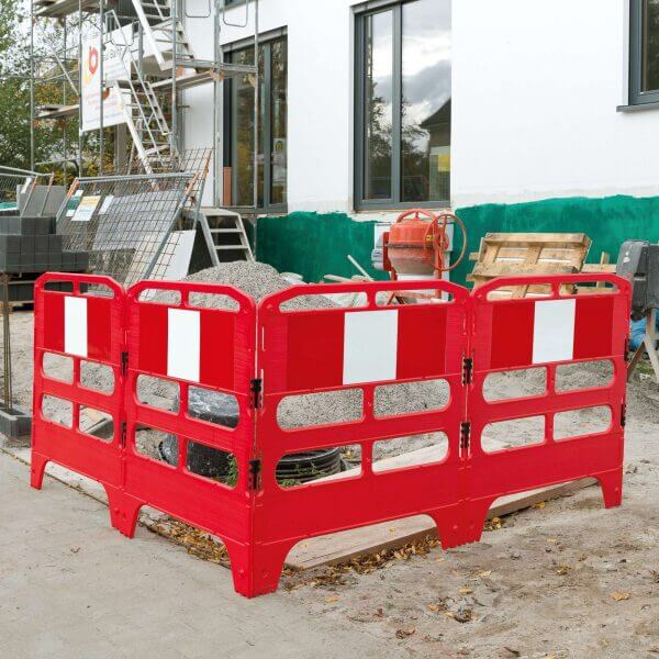 Rote MORION Schachtschutzgitter aus Kunststoff umgeben einen Kieshaufen auf einer Baustelle mit Gerüst und Baumaschinen am Gebäude.