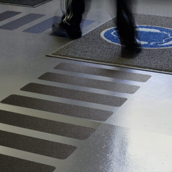 Person mit schwarzer Hose und Schuhen geht über grauen Boden mit PROline Antirutschbelag und rundem blau-grauem Teppich.