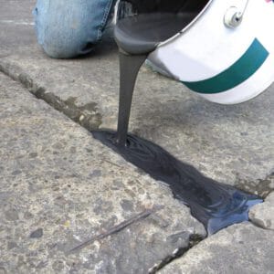 Eine Person gießt Restore Fugenreparatur aus einem Eimer in einen Riss auf einer Betonoberfläche zur Fugensanierung.