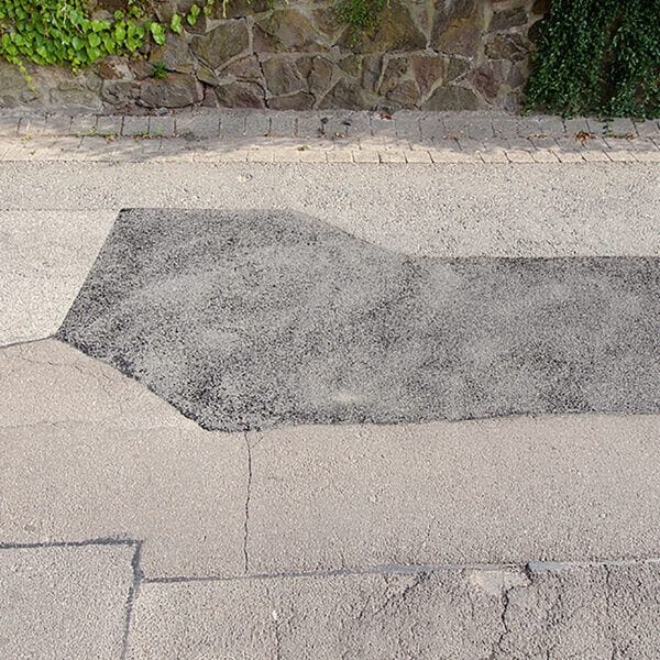 Straßensanierung: Dunkler Asphaltabschnitt hebt sich von älterem, hellem Straßenbelag ab; Steinmauer und Grün im Hintergrund.
