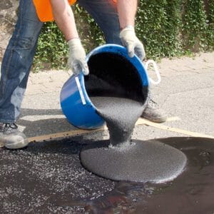 Ein Arbeiter mit Handschuhen und Arbeitskleidung gießt schwarze Asphaltmischung aus blauem Eimer auf Straßenbelag.
