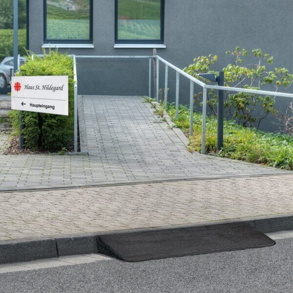 Barrierefreie Betonstrampe mit Metallgeländer, saniert mit Bodenreparatur, führt zum Eingang; Schild weist links zum Haupteingang.