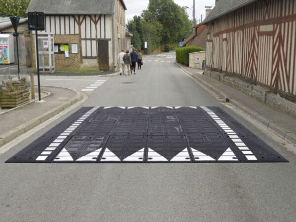 Große schwarz-weiße Bordsteinrampe aus Kautschuk quer über ruhige Wohnstraße, drei Personen gehen im Hintergrund.