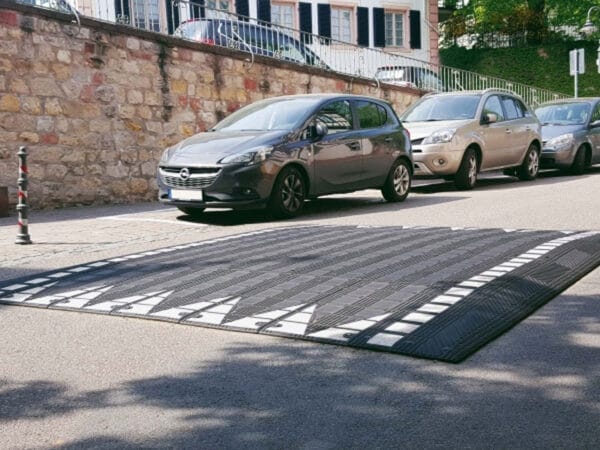 Zwei Autos parken an einer Straße mit Steinmauer; im Vordergrund liegt eine Bordsteinrampe aus Kautschuk auf dem Asphalt.