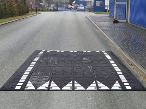 Bordsteinrampe Kautschuk, schwarz-weiß, quer über asphaltierte Straße bei Tor und Industriegebäuden montiert.