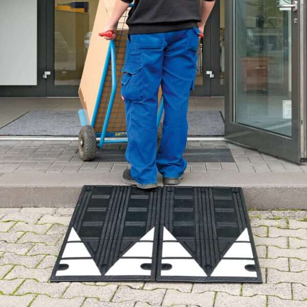 Eine Person in blauer Hose bewegt mit einer Bordsteinrampe Kautschuk und Sackkarre einen großen Karton über den Bordstein am Eingang.