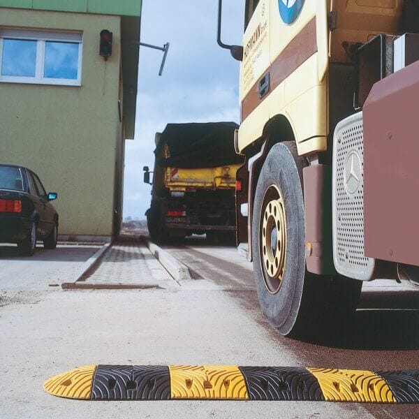 Gelb-schwarze TOPSTOP Fahrbahnschwelle aus Kautschuk auf Straße neben geparktem Lkw und grünem Gebäude montiert.