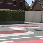 Wohnstraße mit Hecke, weißem Tor, GAMMA Berliner Kissen rot-weiß vor Backsteinhäusern als Verkehrsschwelle.