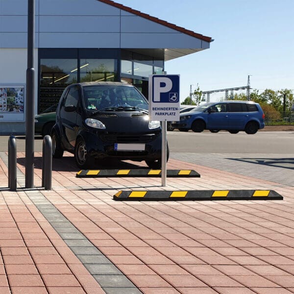Schwarzer Smart mit Radstopp Park-AID auf Behindertenparkplatz vor verglastem Gebäude, blauer PKW im Hintergrund.