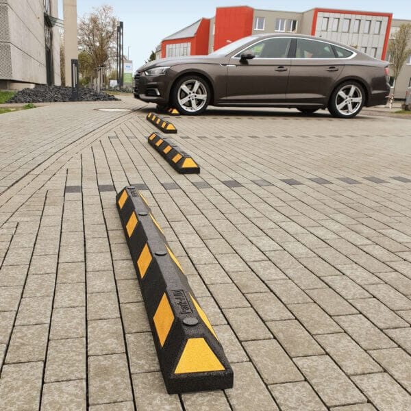 Schwarze-gelbe Radstopp Park-AID Parkplatzbegrenzungen auf Asphalt, braunes Auto, Gebäude im Hintergrund.