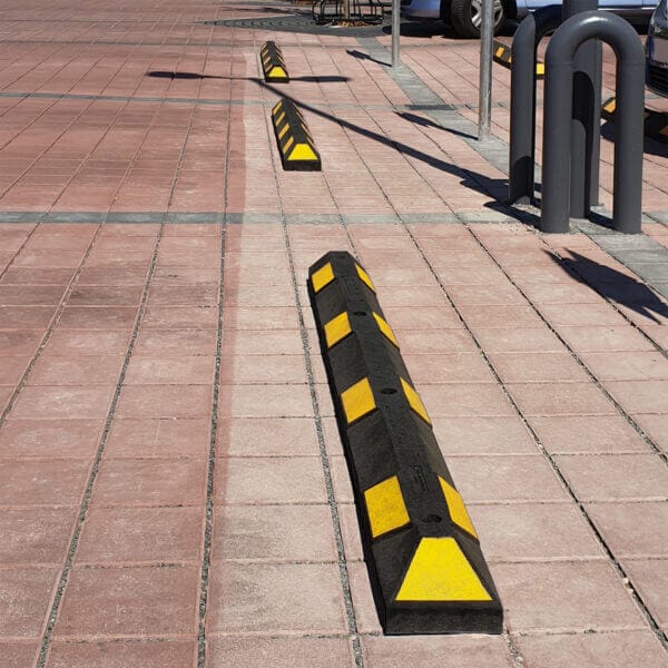 Schwarze und gelbe Radstopper aus Kunststoff auf gepflastertem Parkplatz, mehrere parkende Autos im Hintergrund, Parkplatzmarkierung.