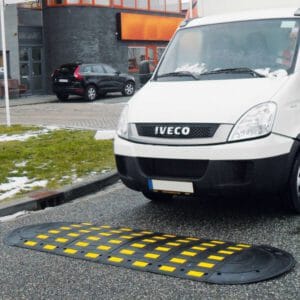 Weißer Lieferwagen nähert sich SafeRide schwarz-gelber Bodenschwelle auf nasser, teils verschneiter Straße vor Gebäude.