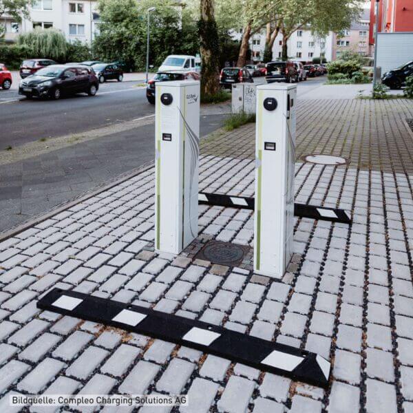 Zwei Radstopp Park-AID Ladestationen für E-Autos auf Gehweg, neben Straße mit geparkten Autos und Mehrfamilienhäusern.