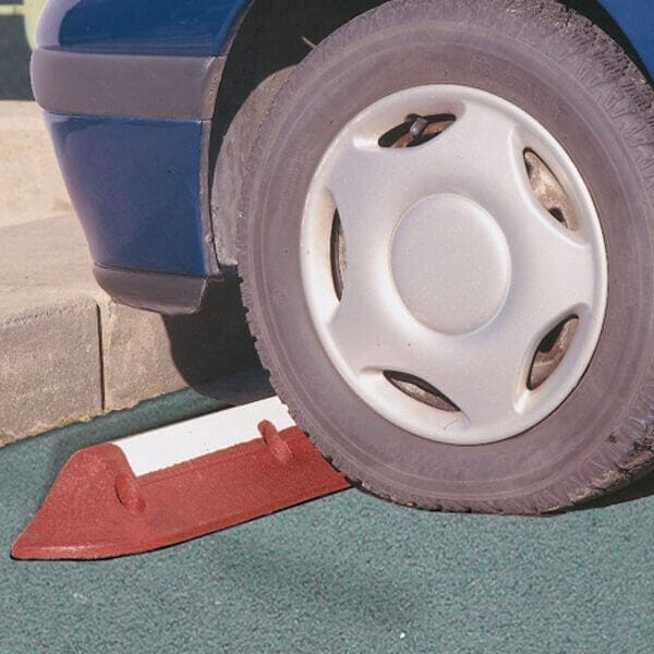 Ein Autoreifen drückt gegen eine rote MORION Radstopp-Barriere auf grün strukturierter Fläche neben einem Betonbordstein.