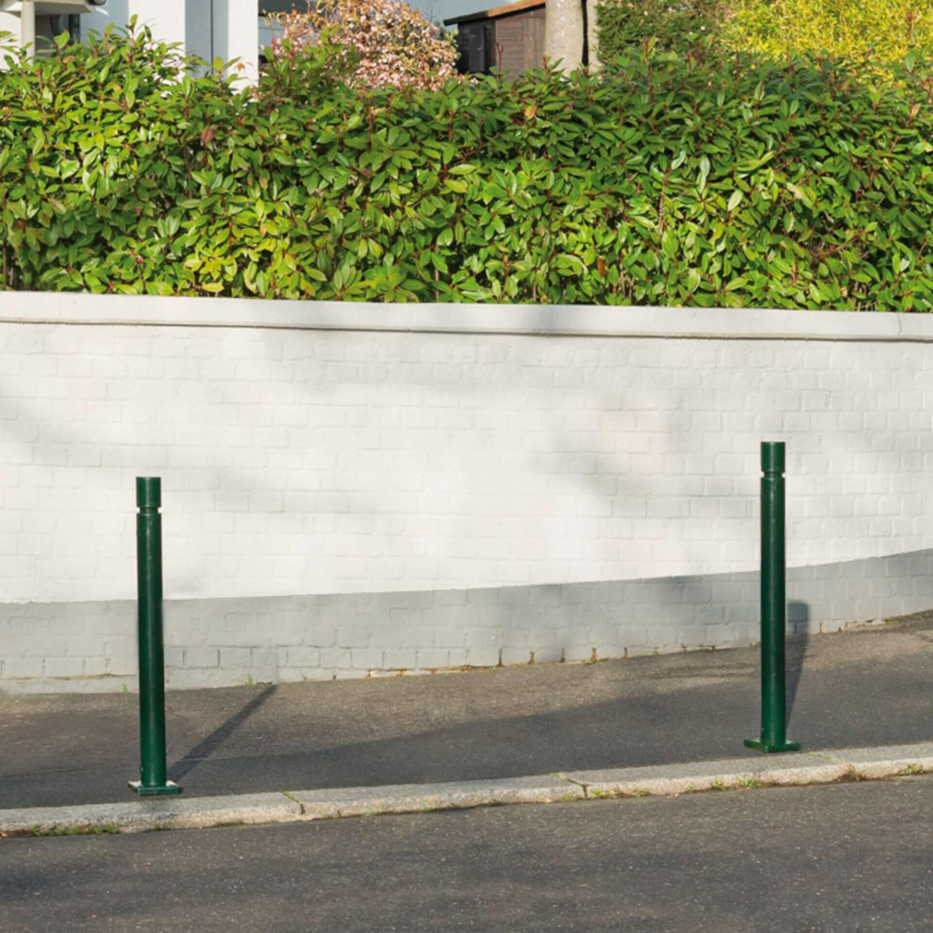 Drei grüne CITY-Poller FLEX aus Metall auf Gehweg vor weißer Backsteinmauer und hoher grüner Hecke in städtischer Umgebung.