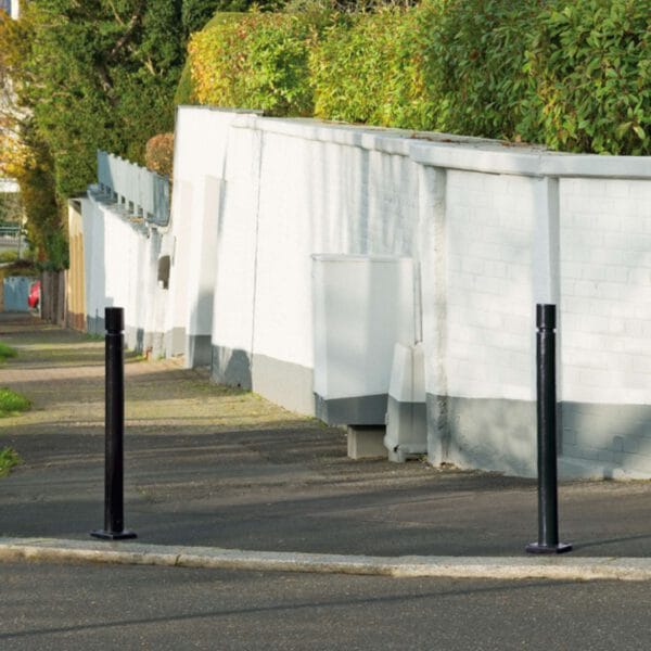 Zwei schwarze CITY-Poller FLEX auf Gehweg am Eingang einer Wohnstraße, dahinter weiße Wand und grüne Büsche.