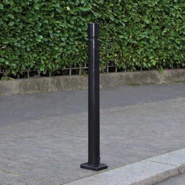 Schwarzer CITY-Poller FLEX aus Metall auf gepflastertem Gehweg am Bordstein, im Hintergrund grüne Hecke.