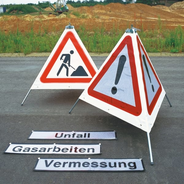 Zwei dreieckige Verkehrsschilder: Warnschild mit Ausrufezeichen, Baustellen-Symbol, daneben deutsche Hinweis-Banner am Boden.