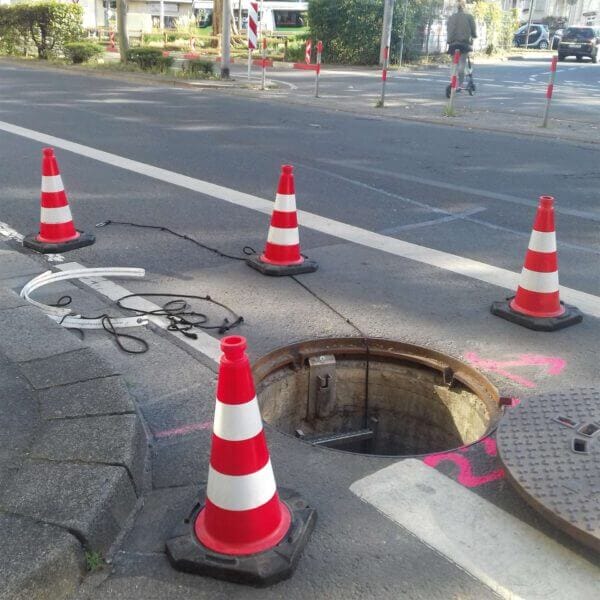 Offener Kanaldeckel auf Stadtstraße, markiert durch MORION ReflexKegel Verkehrsleitkegel, sichtbare Kabel, Markierungen und weitere Kegel.