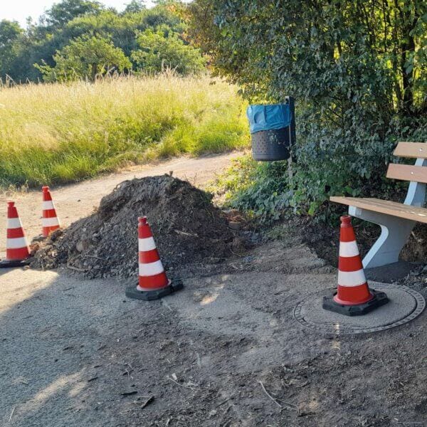 Ein Erdhaufen und vier MORION Reflexkegel EU versperren eine Bank und Mülltonne an einem von Grün umgebenen Gehweg.