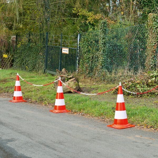 Mehrere tagesleuchtende Leitkegel mit rot-weißen Ketten sperren den Zugang zu einer Wiese an einer asphaltierten Straße vor Privatgrund.