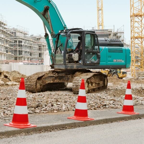 Baustelle mit blauem Bagger, drei MORION-Leitkegel-EU orange-weiß, Kiesboden, unfertige Gebäude im Hintergrund.
