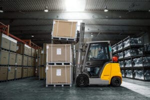 Ein Gabelstapler transportiert große Kartons in einem Lager mit Palettenstapeln und Regalen aus Metall.