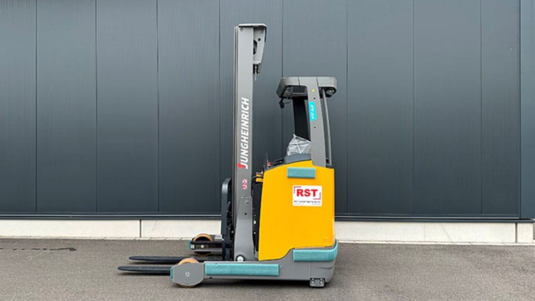 Gelber Jungheinrich Elektro-Schubmaststapler steht auf Asphalt vor dunkler, grauer Industriehalle.